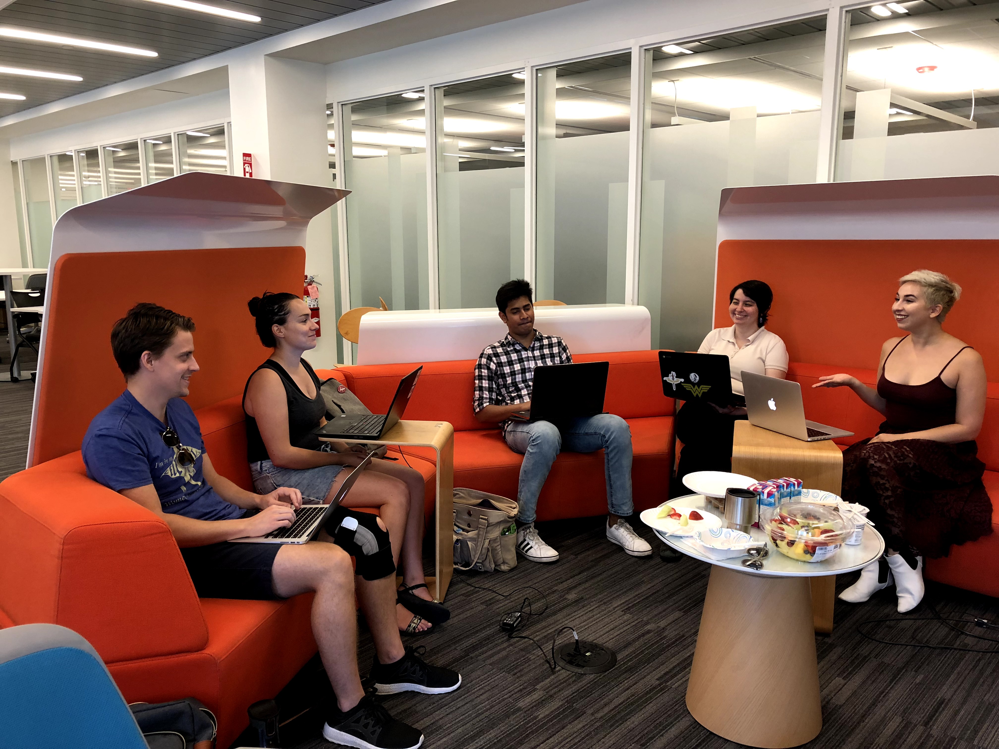 Five people sit on a couch in a semi-circle, holding laptops. They are talking.